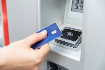 A person holding a blue contactless credit card near an ATM reader to make a tap payment, illustrating modern banking technology and secure contactless transactions.