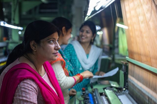 Three individuals in an industrial textile facility examining machinery and discussing operations, with one holding a document, highlighting teamwork and manufacturing processes.