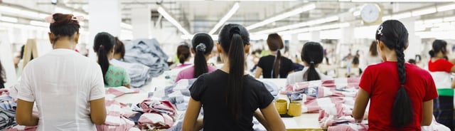 Workers in a garment factory sorting and folding fabric on large tables under bright overhead lights.
